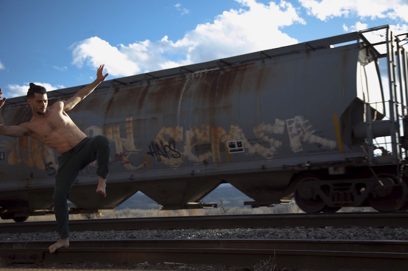 A man, shirtless and barefoot, balances on train tracks. An old train is behind him.